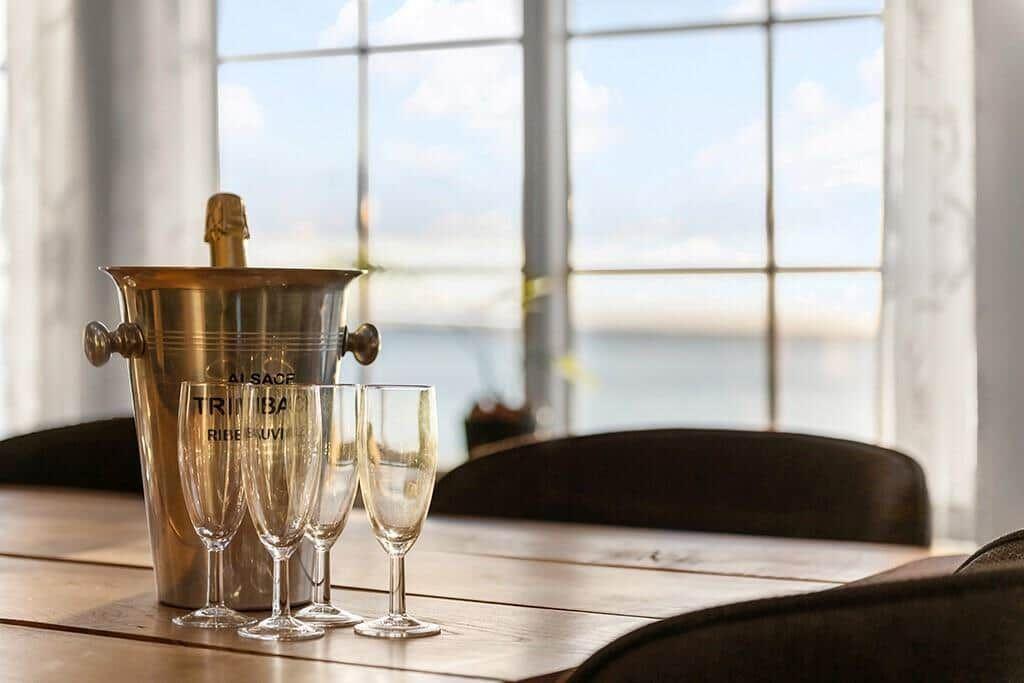 On the table are glasses and an ice bucket with champagne. Water is visible through the window.