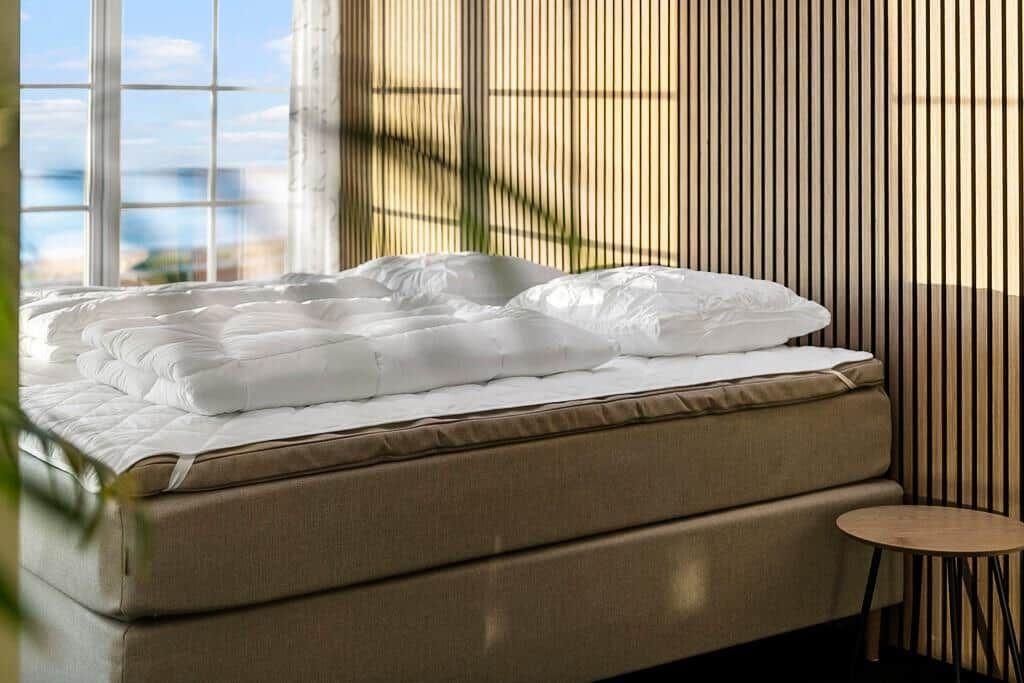 A bed with white pillows and a down comforter. Behind the bed, a window with a view of the sea.