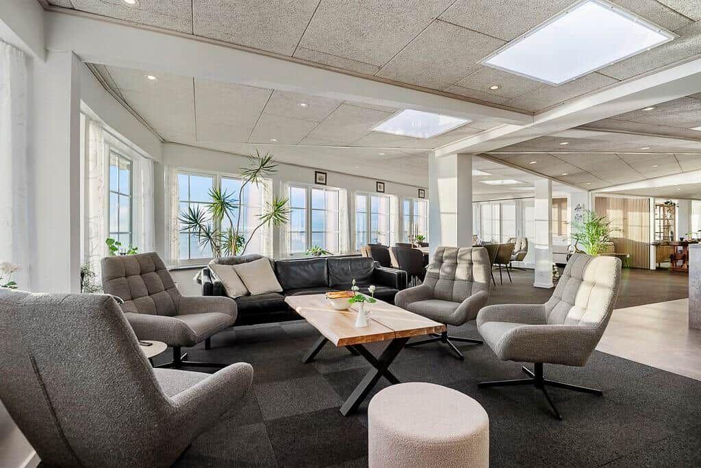 Large living area with seating, table, and plants. Large windows and light ceiling.