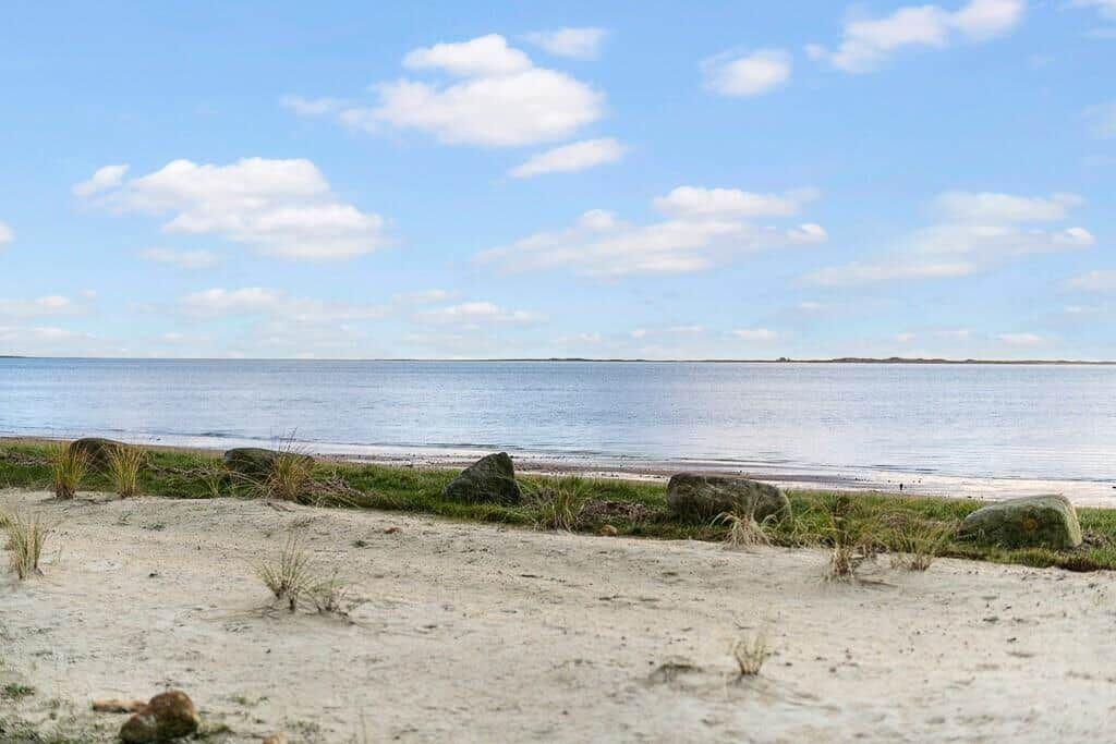 Beach with sand, grass, and stones by the water under a blue sky with white clouds.