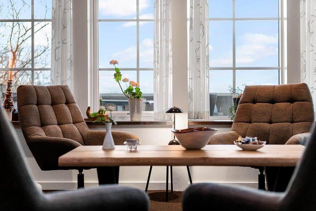 Two brown armchairs surround a wooden table. Large windows overlook the sky.