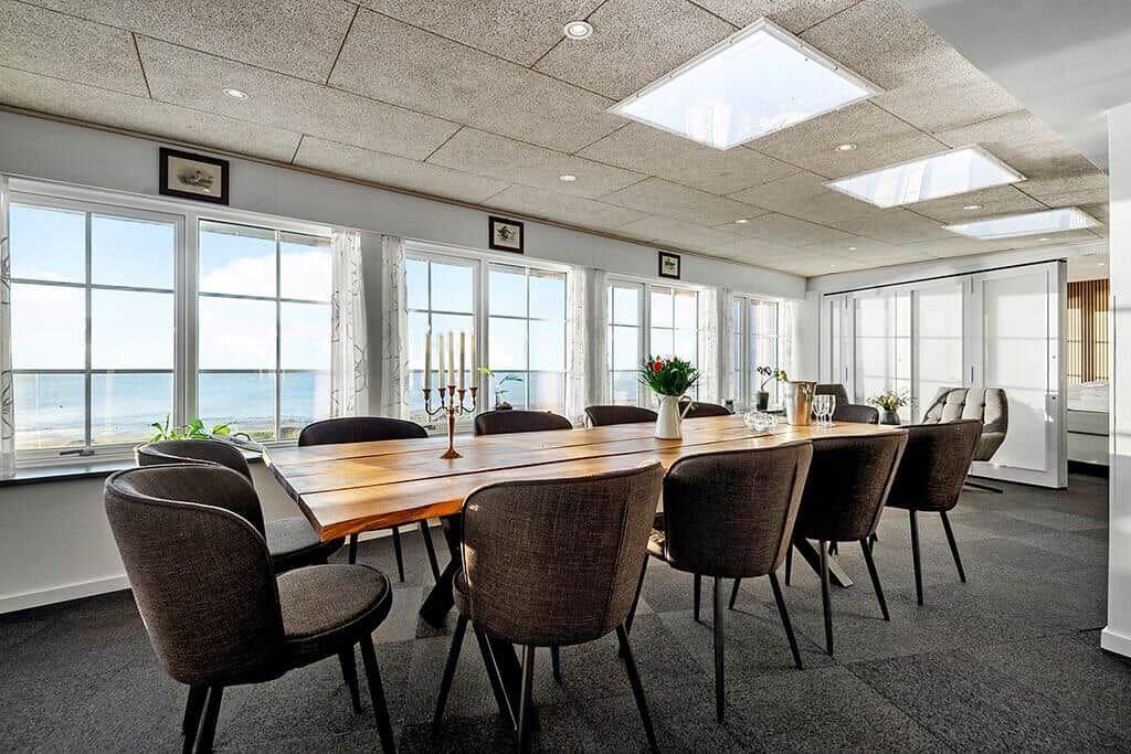 Dining room with long wooden table, chairs, and sea view through large windows.