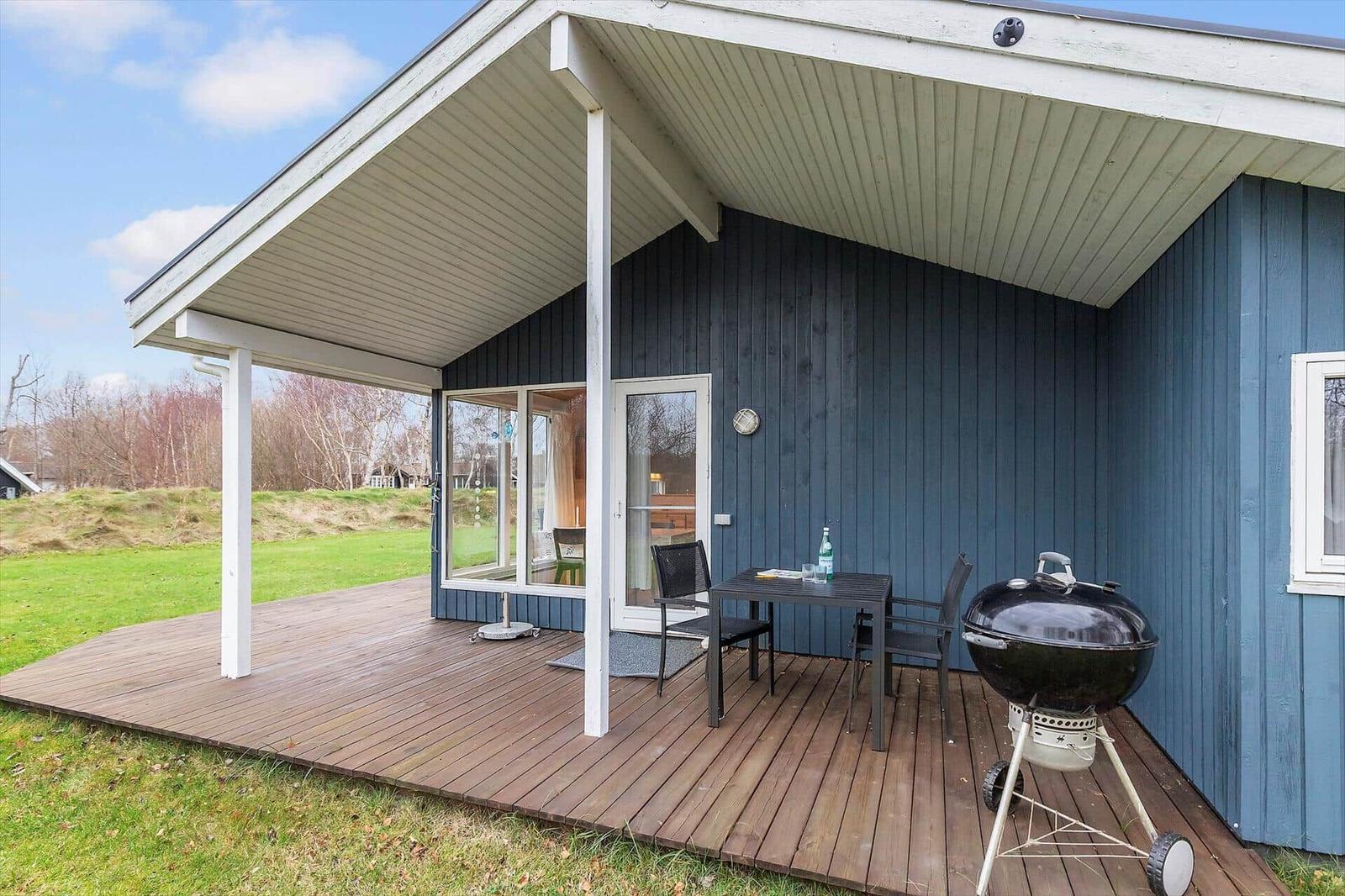 Deck with table, chairs, and grill in front of blue wooden house with glass doors.