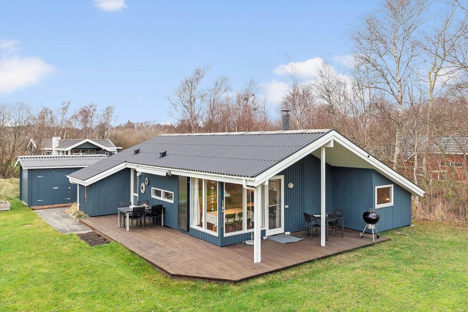 A blue holiday home with wooden terrace, grill and garden. Large windows and gable roof.