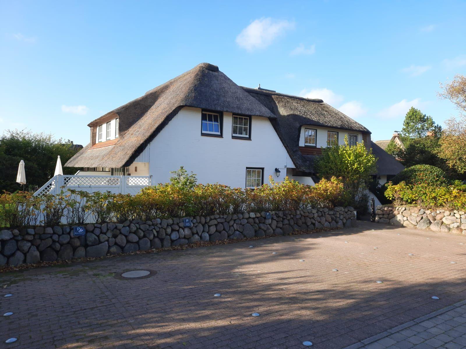 Haus mit strohgedecktem Dach, weiß verputzt, umgeben von Steinmauer und Bäumen.