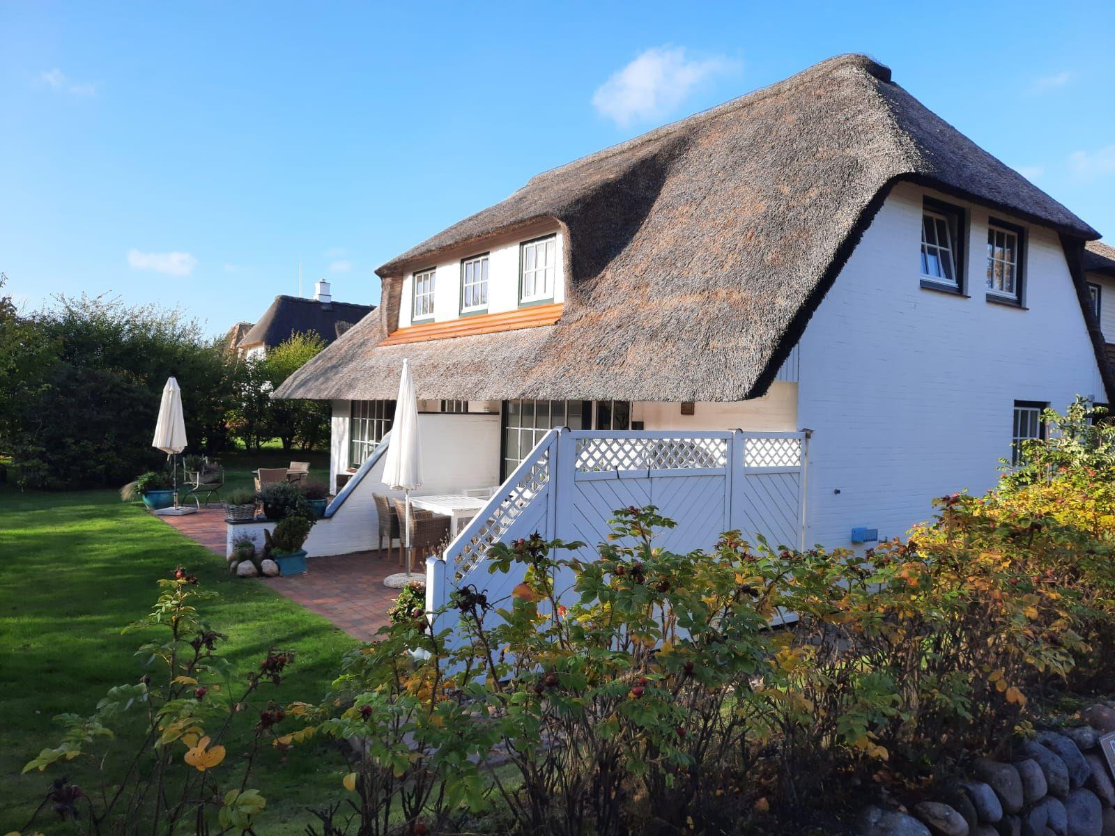 Ein weißes Haus mit Strohdach, Garten und Terrasse mit Gartenmöbeln.