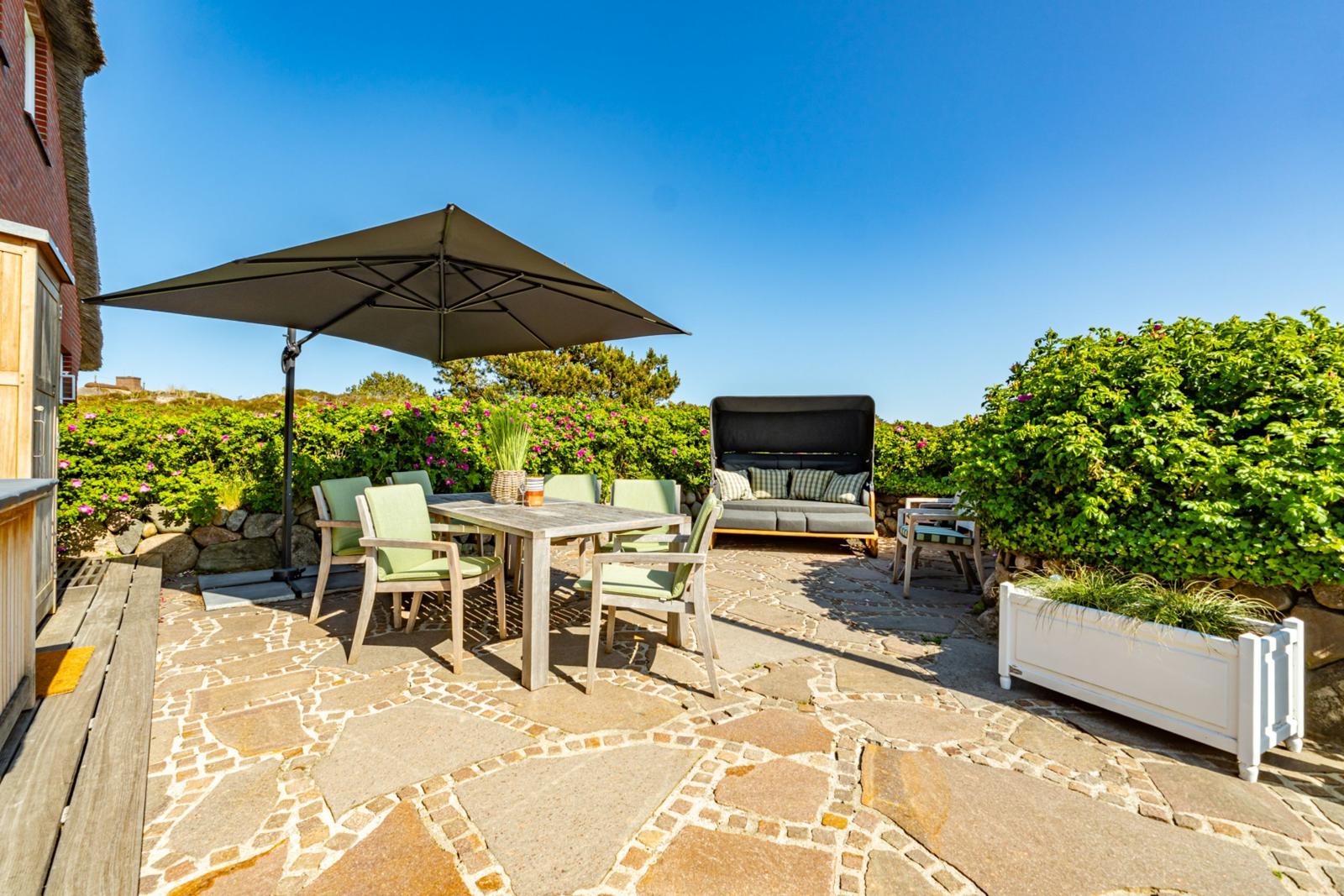 Terrasse mit Tisch, Stühlen, Sonnenschirm und Liege unter blauem Himmel.