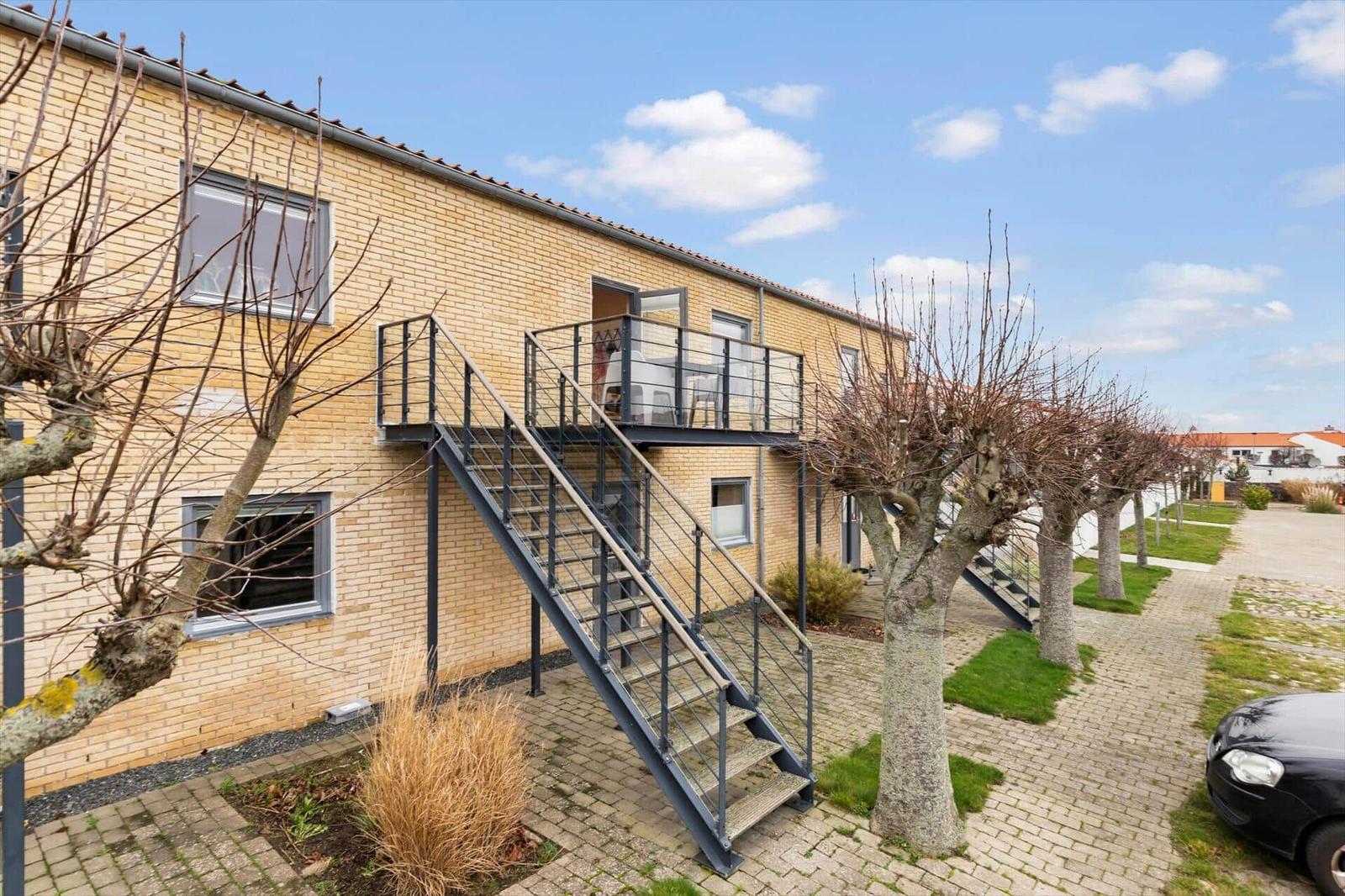 A two-story house with balcony and metal stairs. Trees and paved paths.