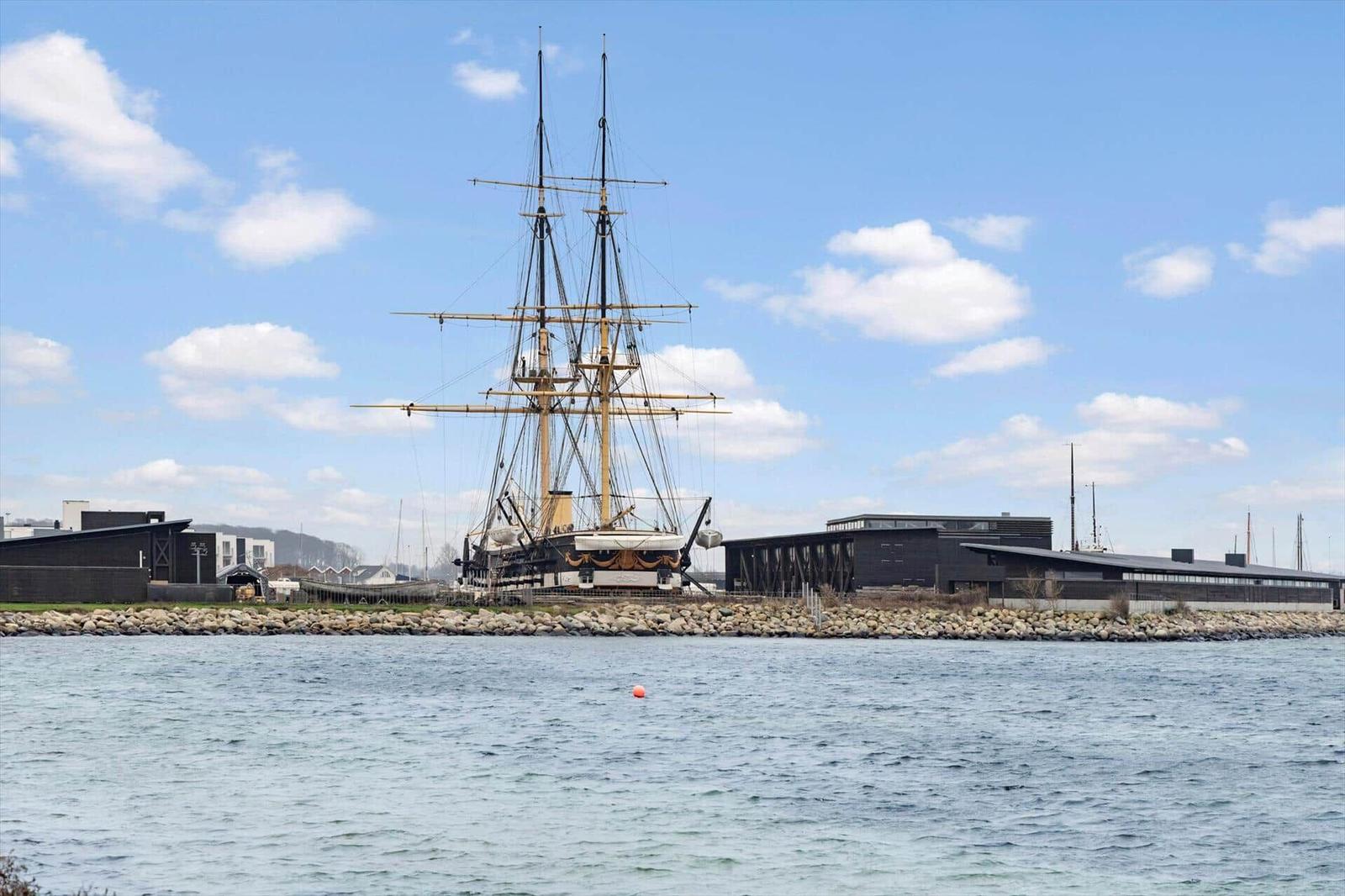 Historic sailing ship in harbor with modern buildings in background.