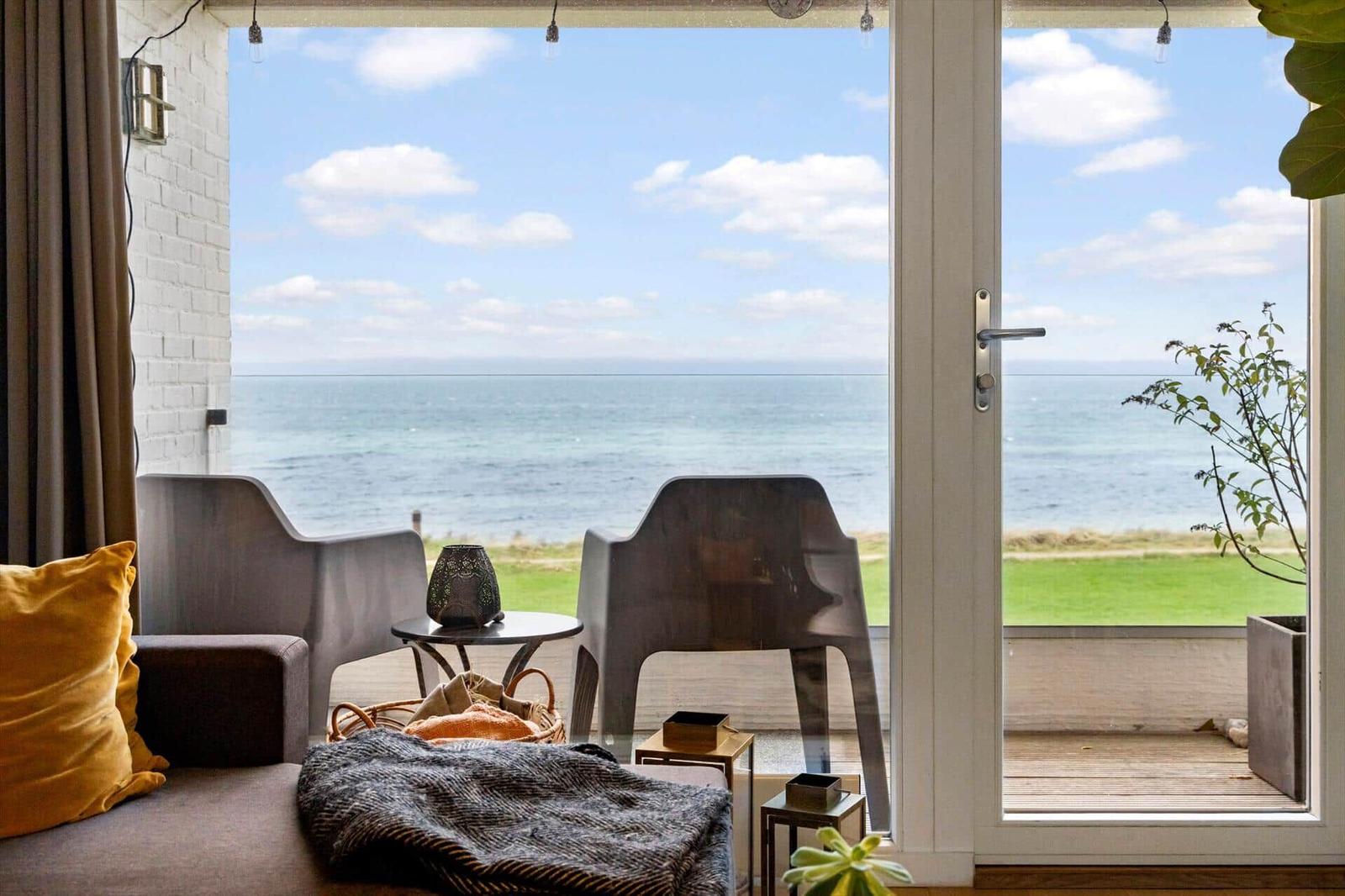 Through the glass door, you see the sea. Inside, there is a sofa with cushions.