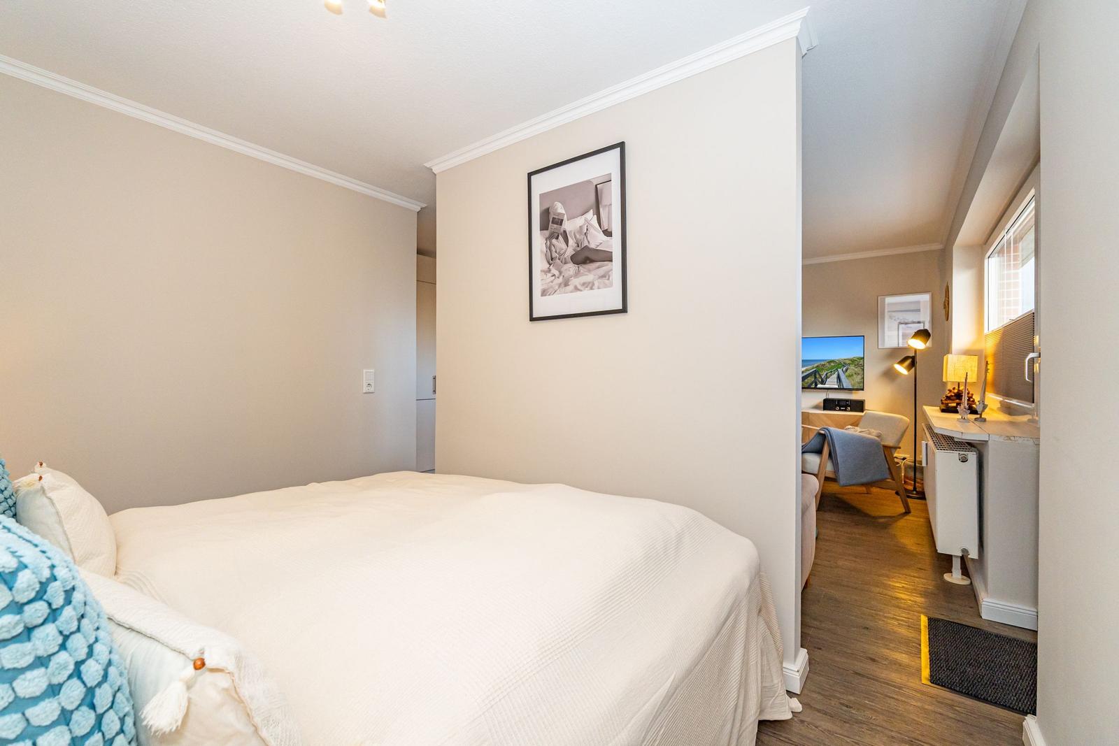 Schlafzimmer mit Bett und Bild an der Wand. Durchgang zu Wohnbereich mit Fernseher.