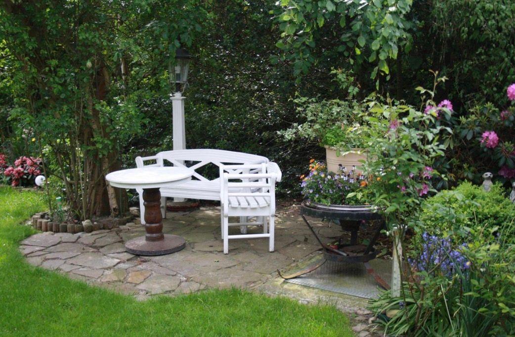 Weißer Sitzbereich mit Tisch auf Steinplatten im Garten mit Pflanzen und Beleuchtung.