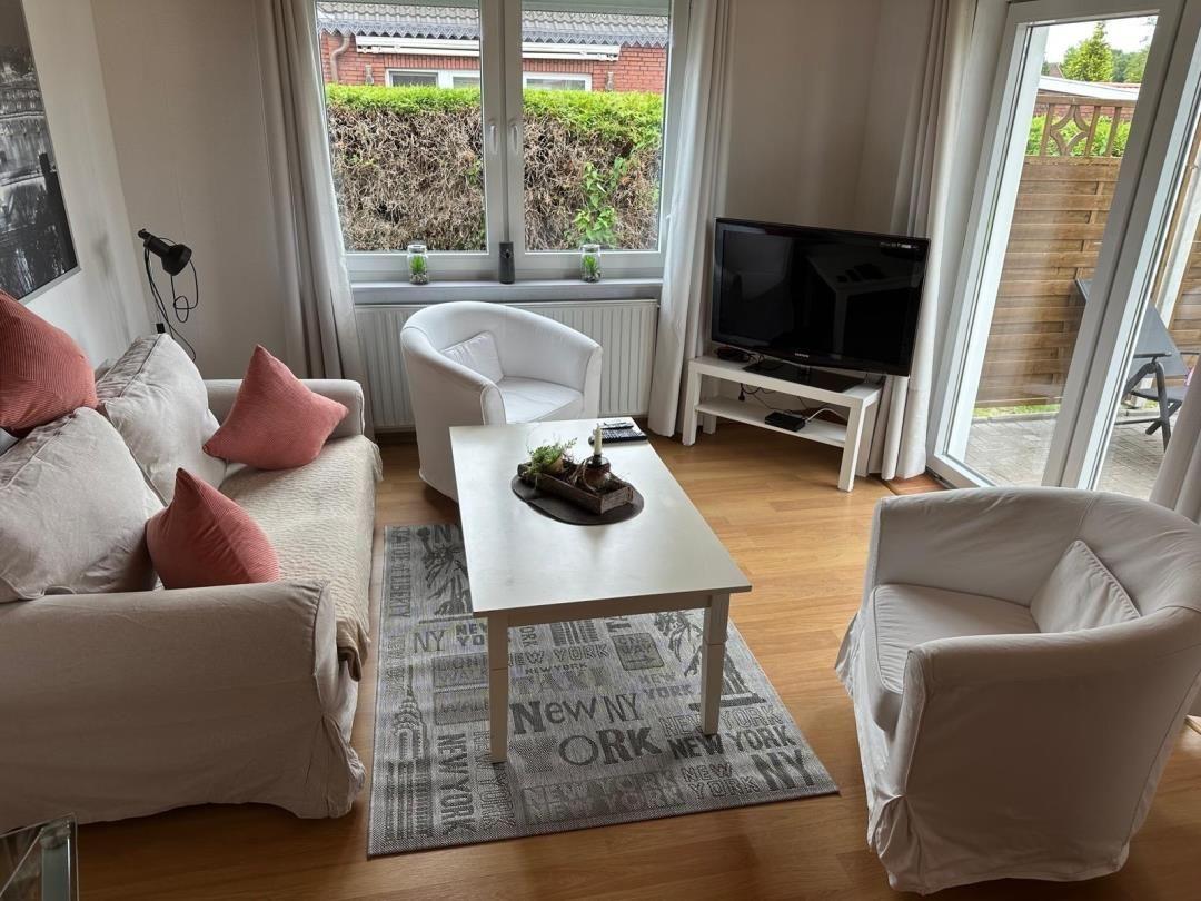 Wohnzimmer mit Sofa, zwei Sesseln, Couchtisch und Fernseher. Durchgang zur Terrasse.