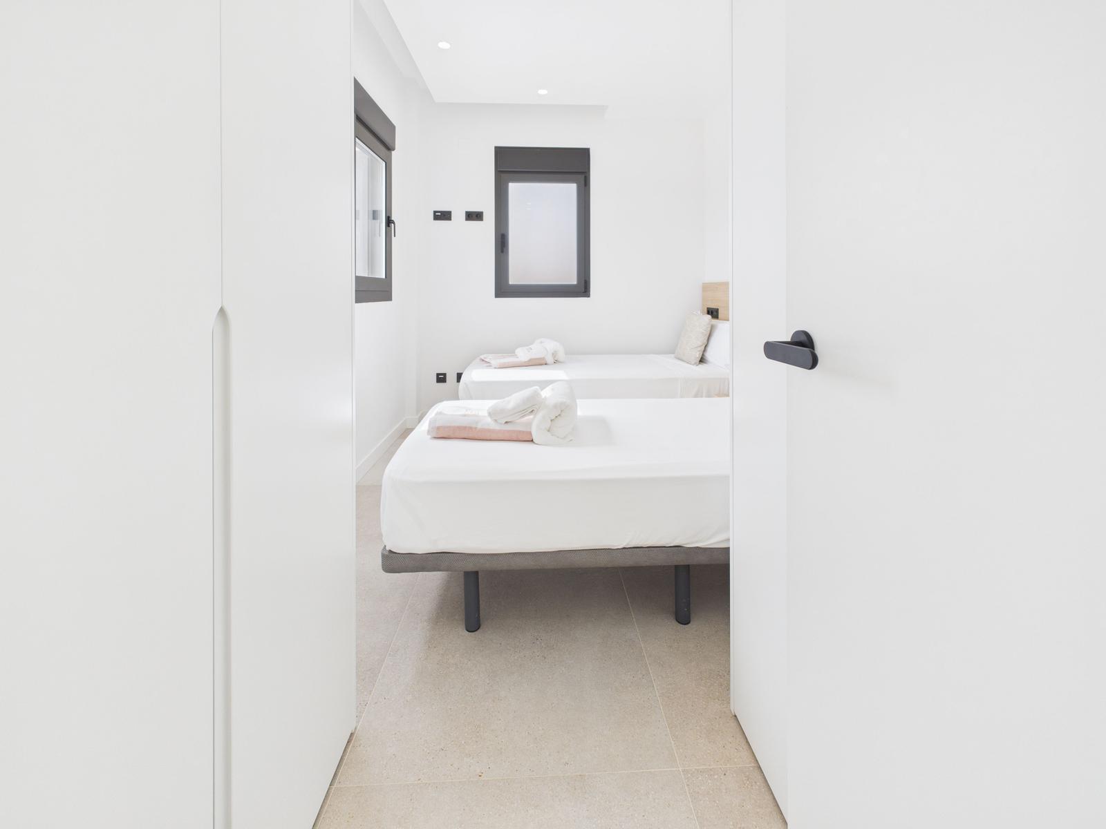 Two beds with white sheets and towels. White walls and dark window frames.