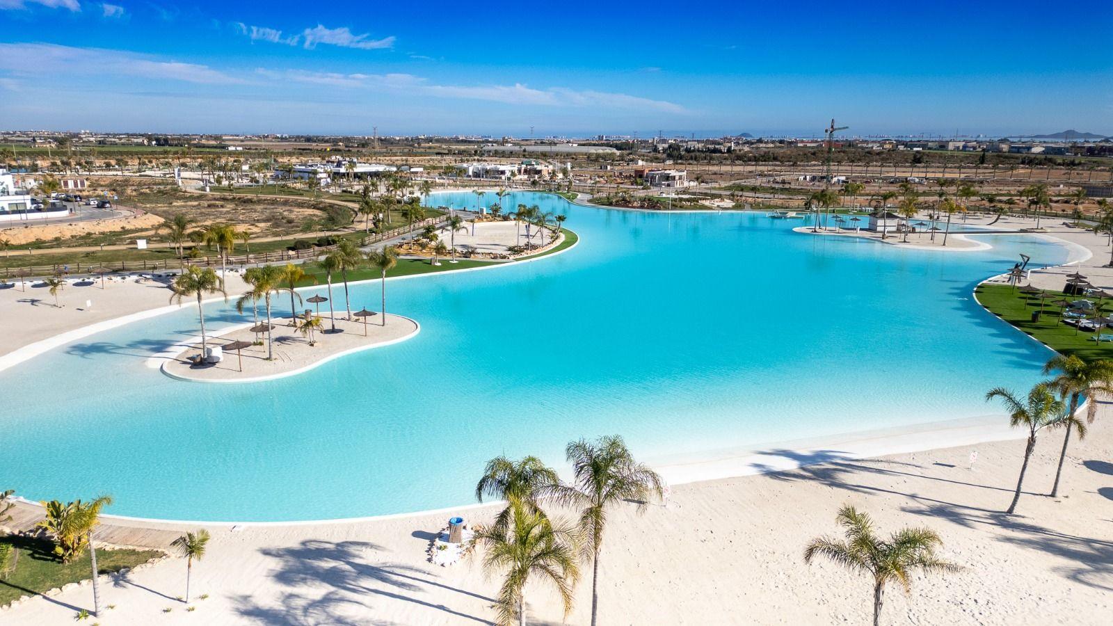 Large pool with sandy areas and palm trees under blue sky.