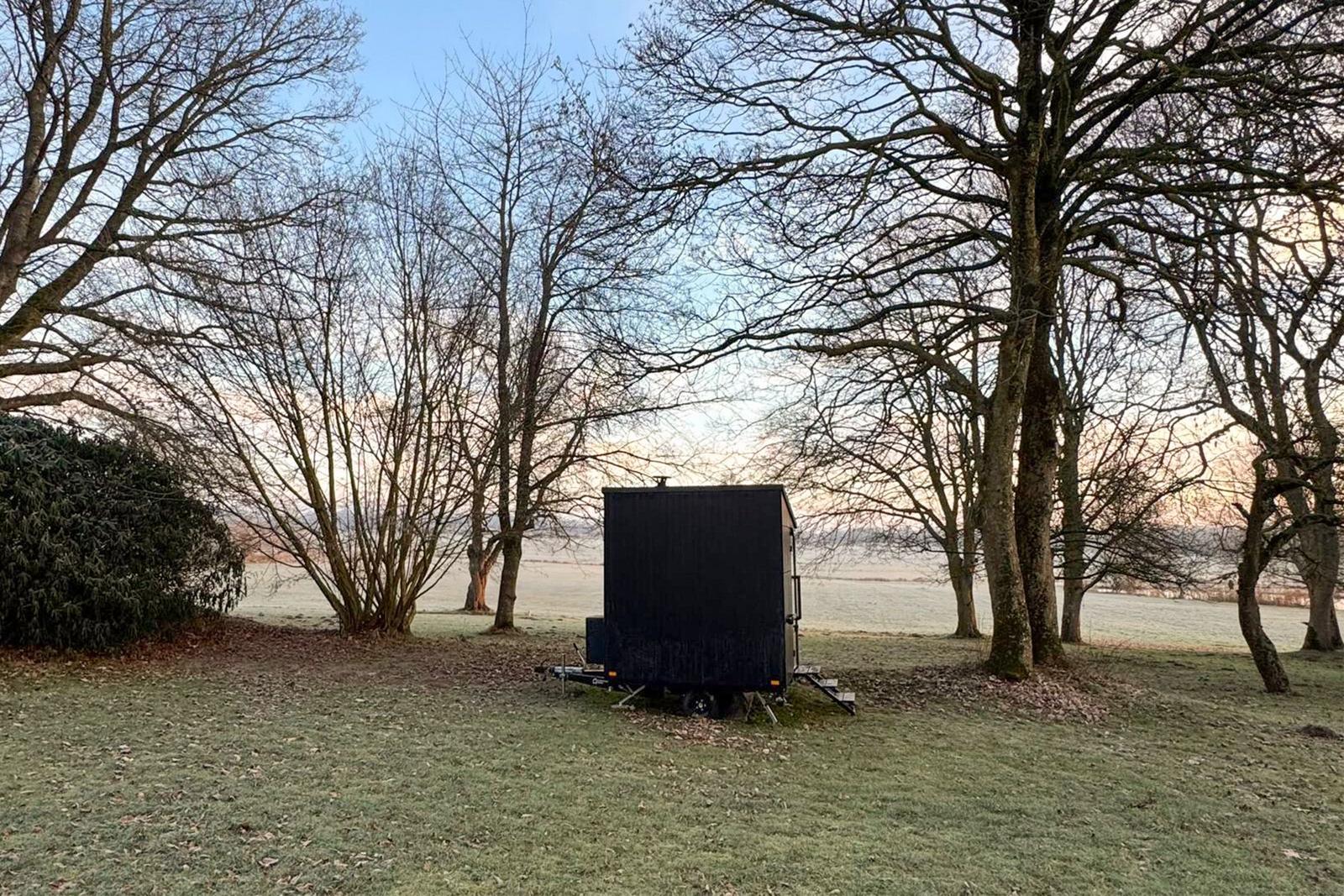 Black trailer on grass field between bare trees with view of field.