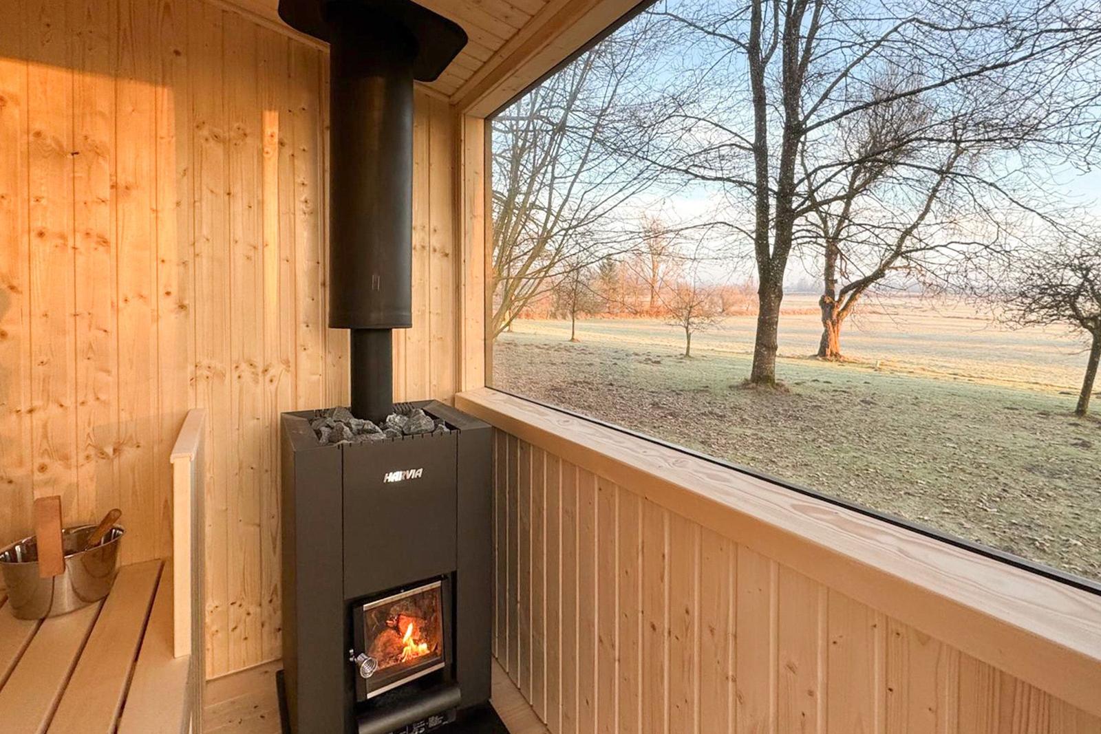 Sauna with wooden walls and wood-burning stove, view of field and trees.