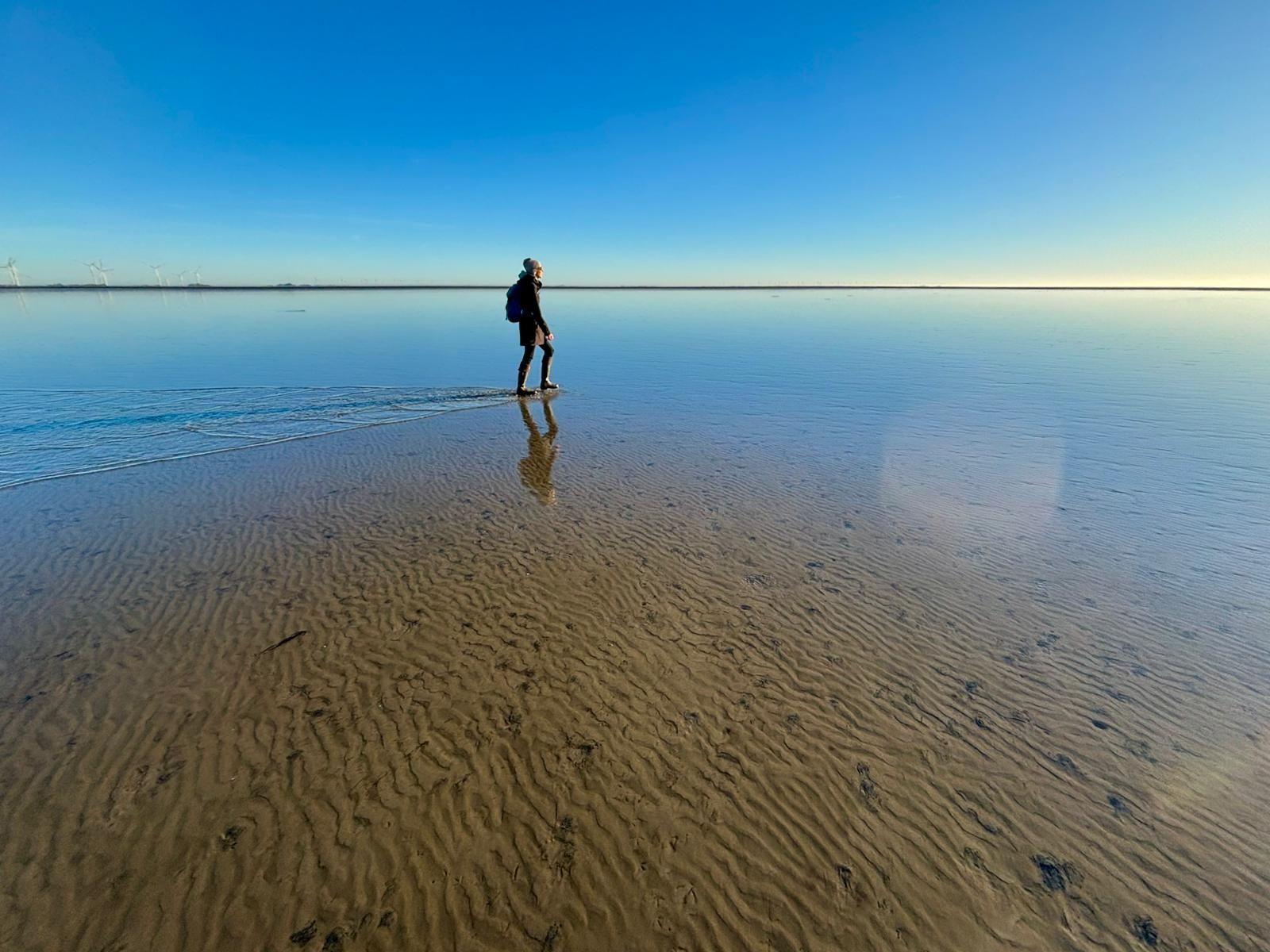 Eine Person geht durch flaches Wasser auf einem sandigen Strand. Windräder sind im Hintergrund sichtbar.