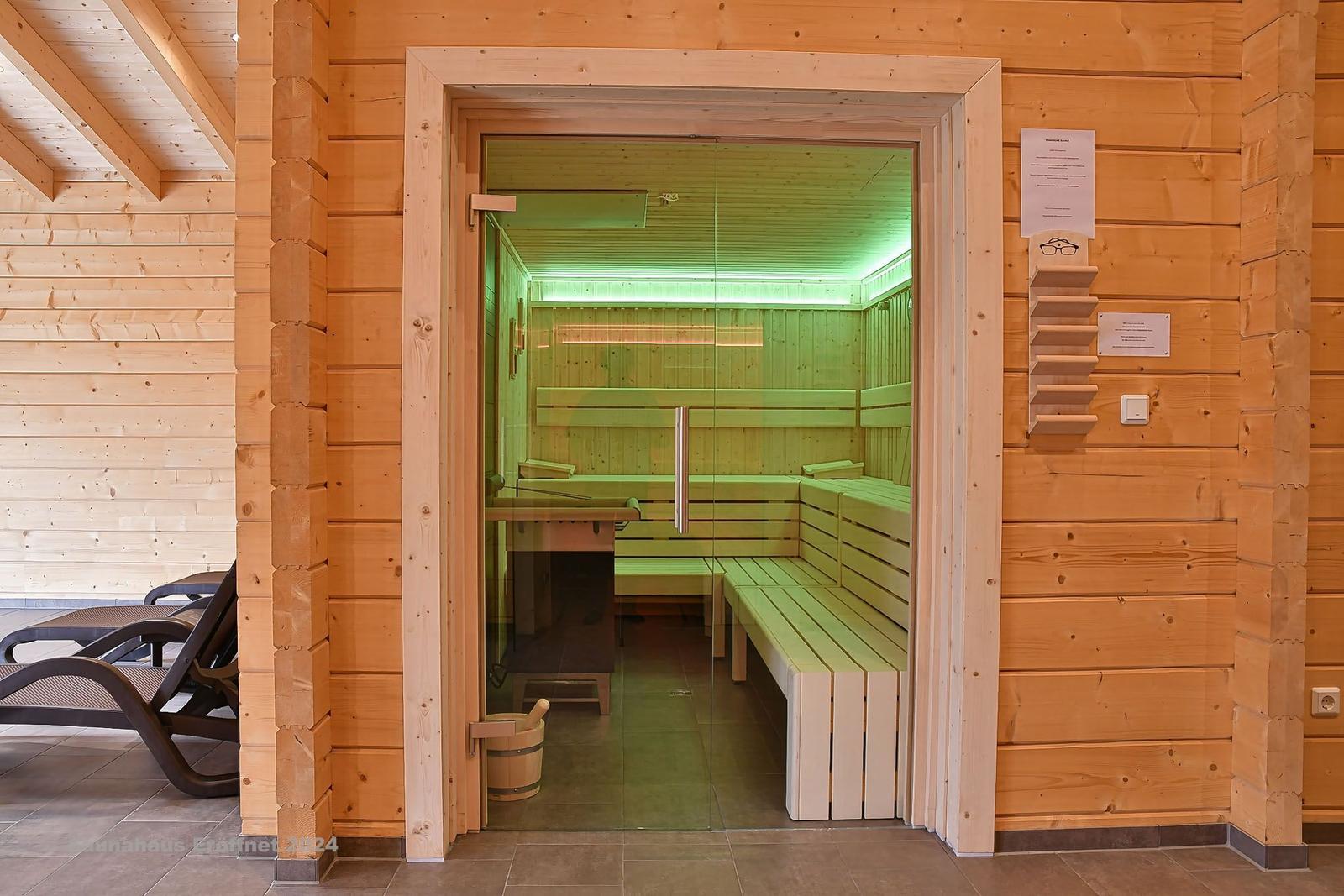 Sauna mit Holzbänken und Glaswand. Links Liegestühle. Rechts Wand mit Ablage und Schalter.