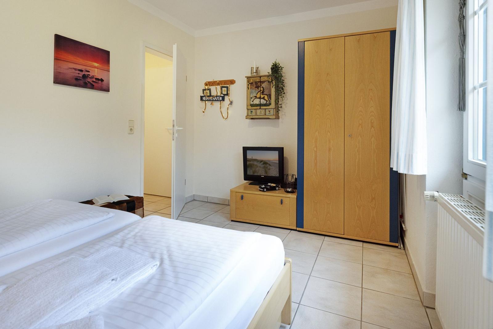 Zimmer mit Doppelbett, Kleiderschrank, TV und Fenster mit Vorhang.