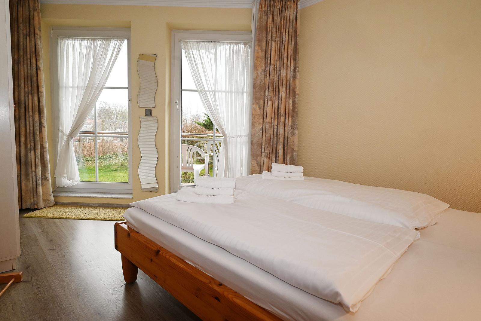 Schlafzimmer mit Doppelbett, zwei Fenstern und Blick auf Balkon.