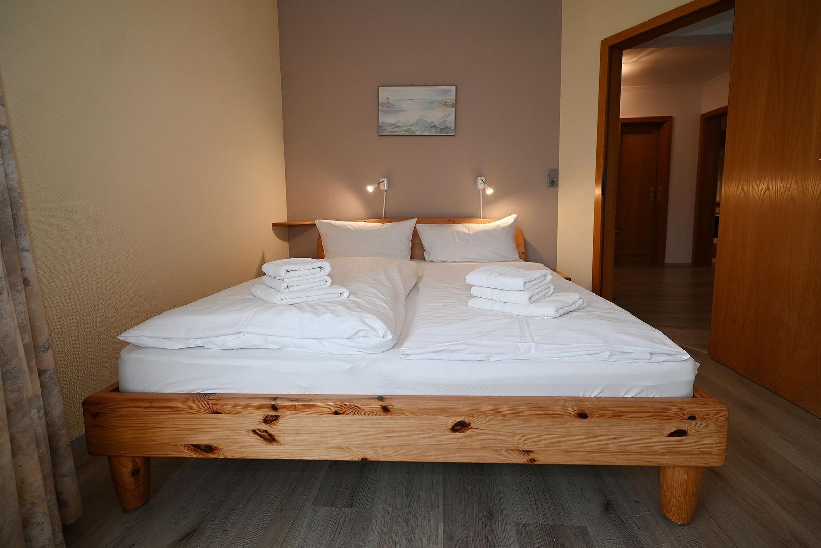 Ein Bett mit weißer Bettwäsche und gefalteten Handtüchern. Holzrahmen und Wandbilder.