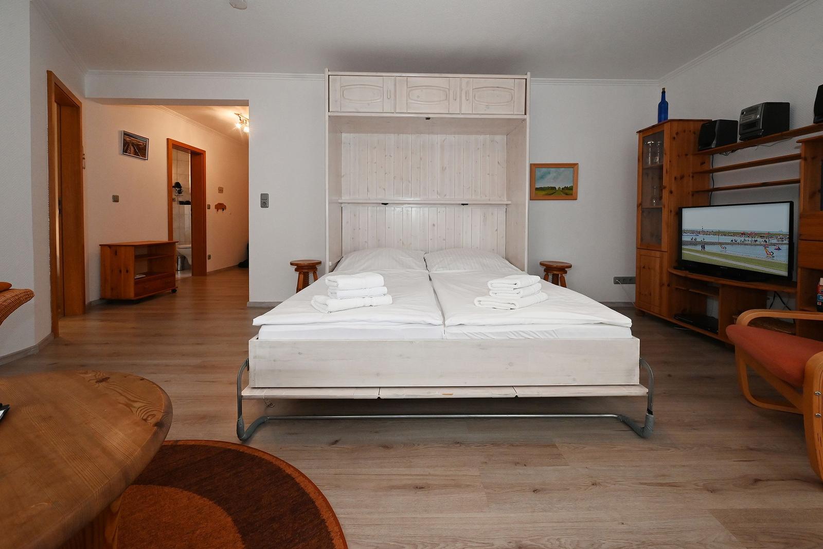 Schlafzimmer mit Bett, Fernseher und Holzmöbel. Durchgang zur Badewanne.