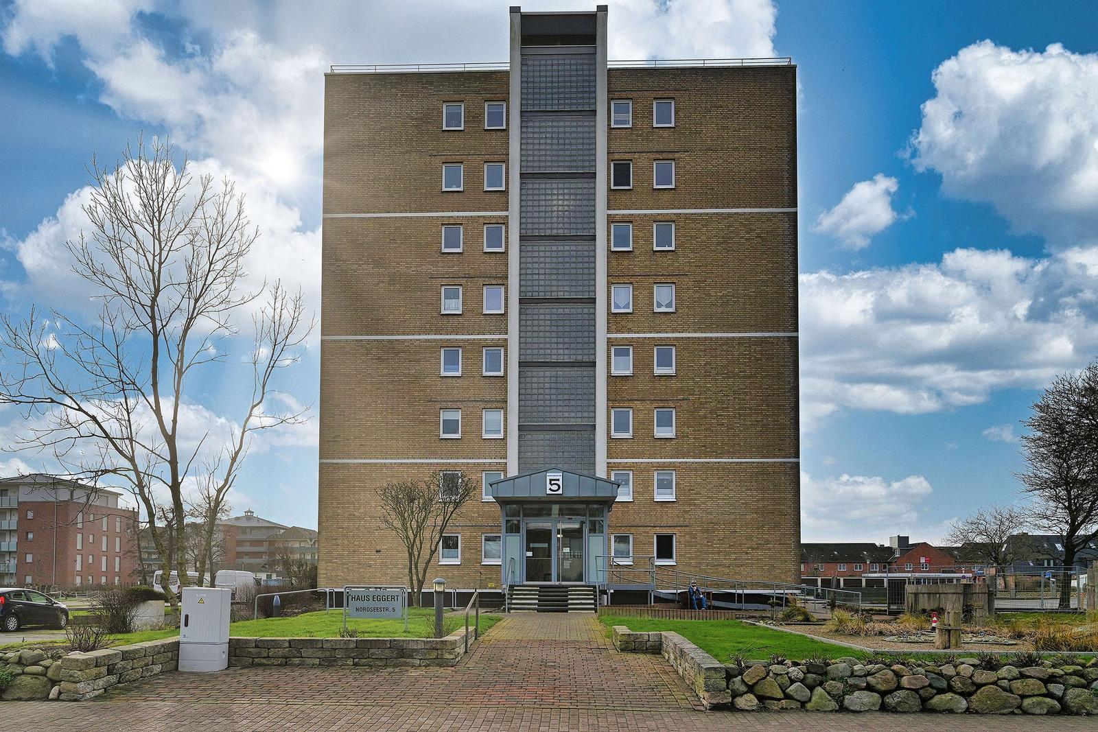 Hochhaus mit Eingang und Grünanlagen unter blauem Himmel.