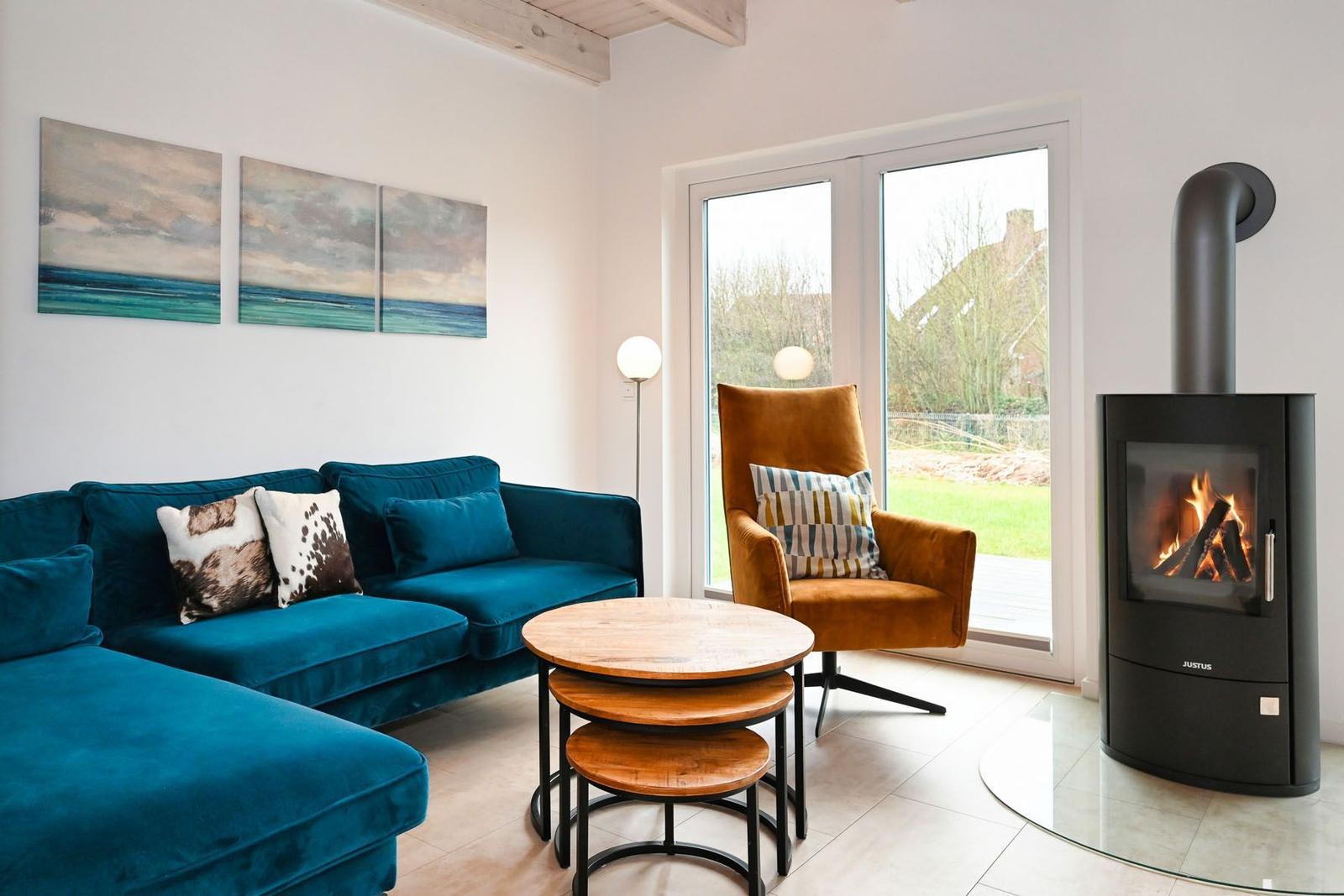 Ein Wohnzimmer mit blauem Sofa, orangefarbenem Sessel und Kaminofen. Große Fenster und Wandbilder.