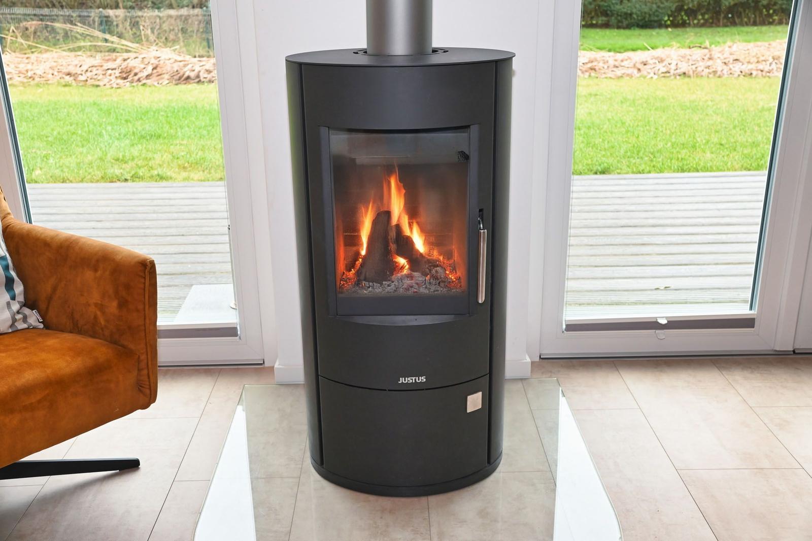 Ein Kaminofen mit brennendem Holz steht vor einer Glasfront. Ein brauner Sessel ist links im Bild.