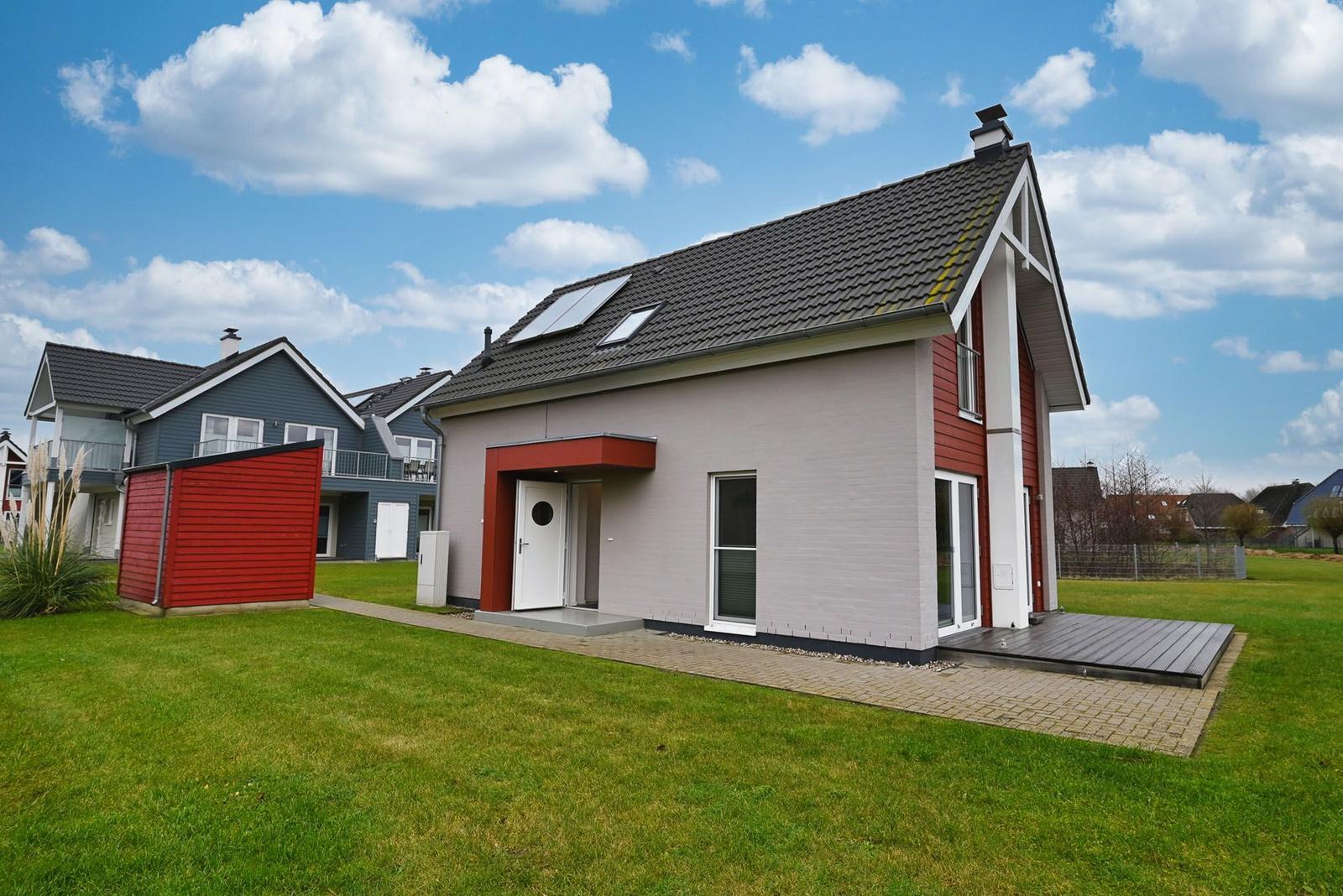 Moderne Villa mit rotem Anbau, Garten und Terrasse unter blauem Himmel.