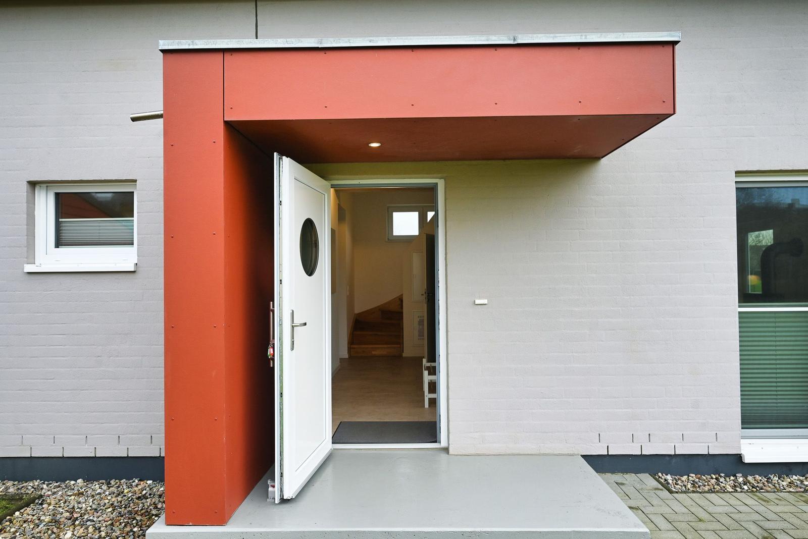 Haus mit rotem Vordach und weißer Eingangstür. Durchgang ins Innere mit Holztreppe.