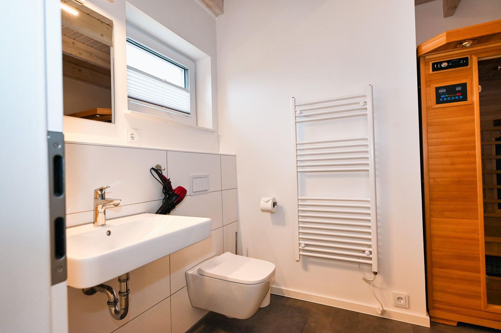 Badezimmer mit Waschbecken, Toilette und Heizkörper. Nebenraum mit Holzsauna.