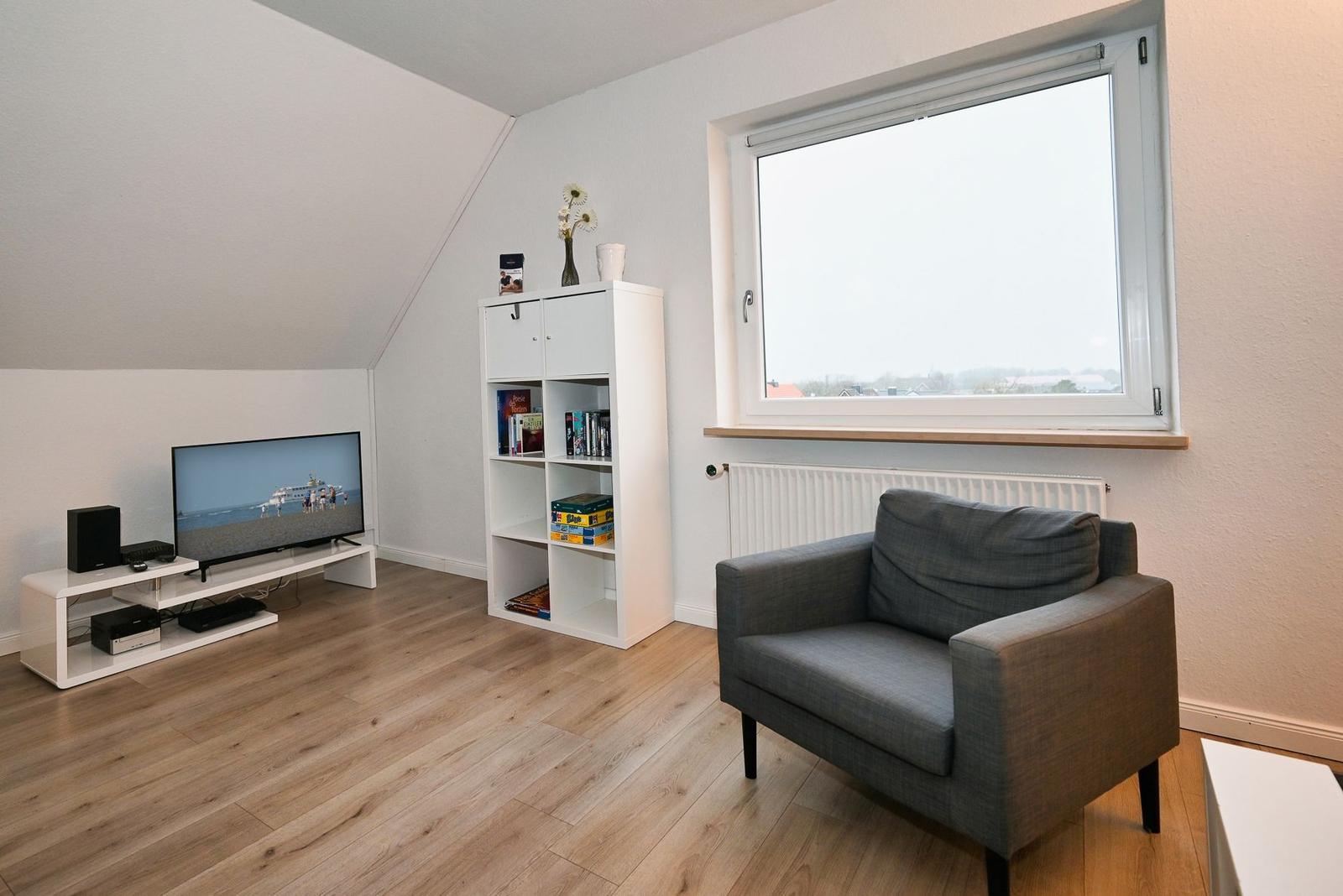 Wohnzimmer mit grauem Sessel, TV und Bücherregal. Holzboden und großes Fenster.