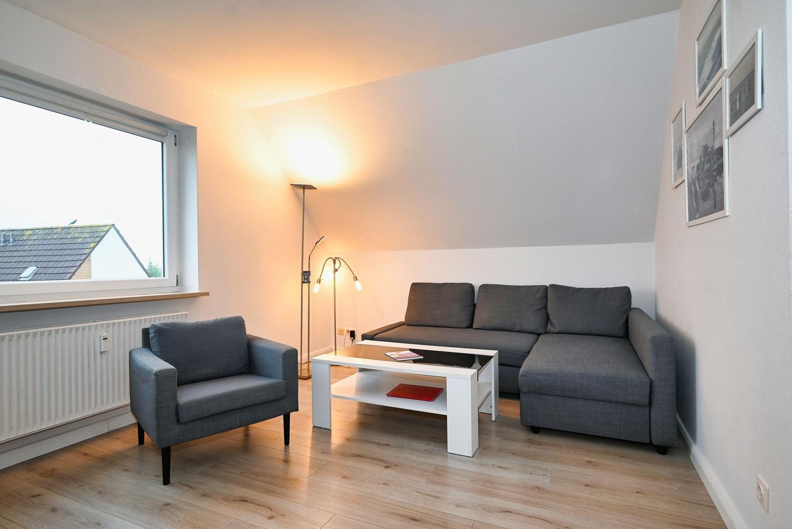 Wohnzimmer mit grauem Sofa, Sessel und Tisch. Fenster und Wandbilder sind sichtbar.