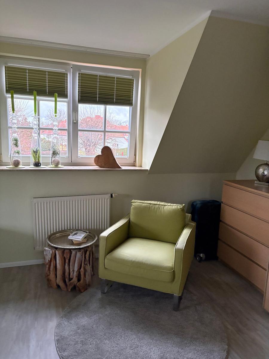 Ein Zimmer mit grünem Sessel, Holzschrank und Fenster mit Jalousien.