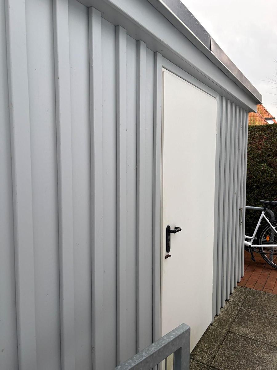 Graue Metallgarage mit weißer Tür und schwarzer Klinke. Fahrrad steht daneben.