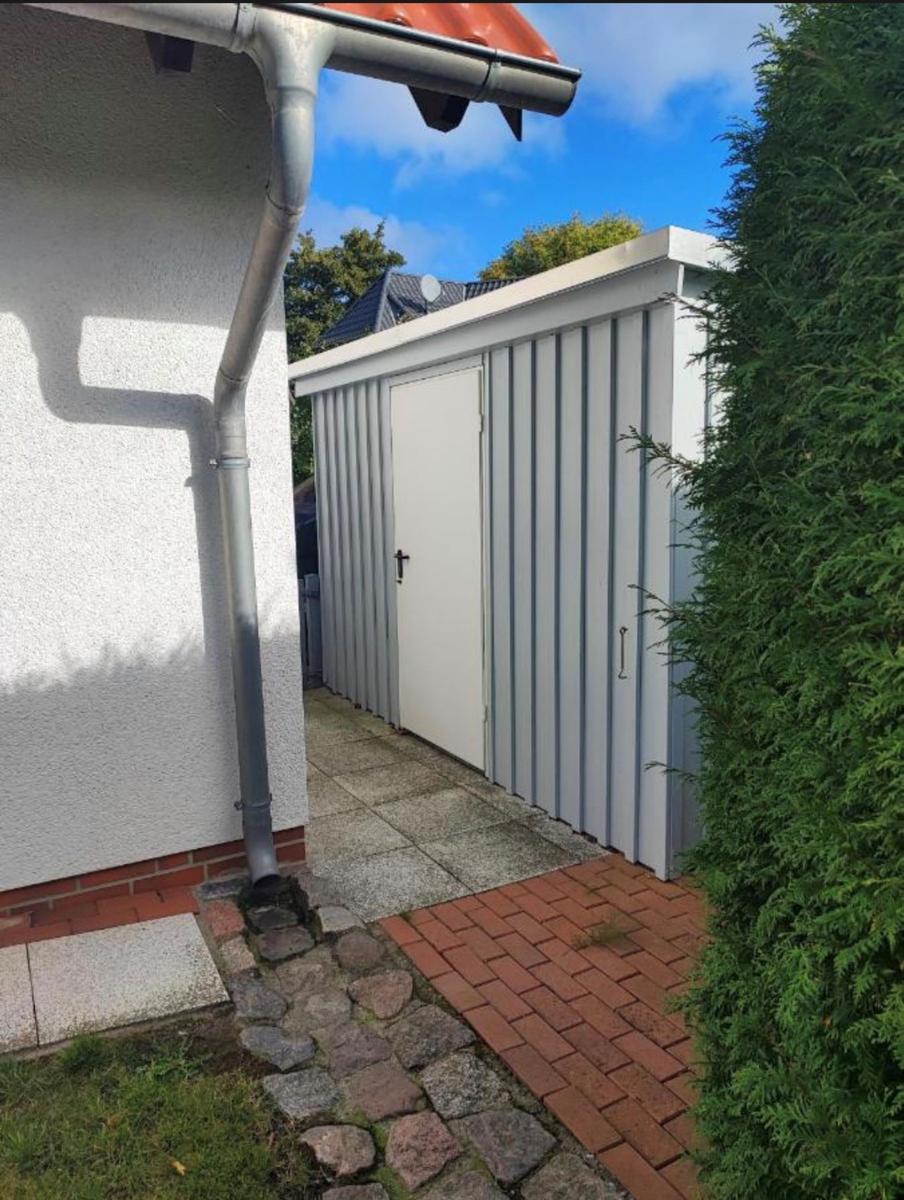 Ein weißer Metallschuppen mit Tür neben einem Haus und einem Ziergehölz.