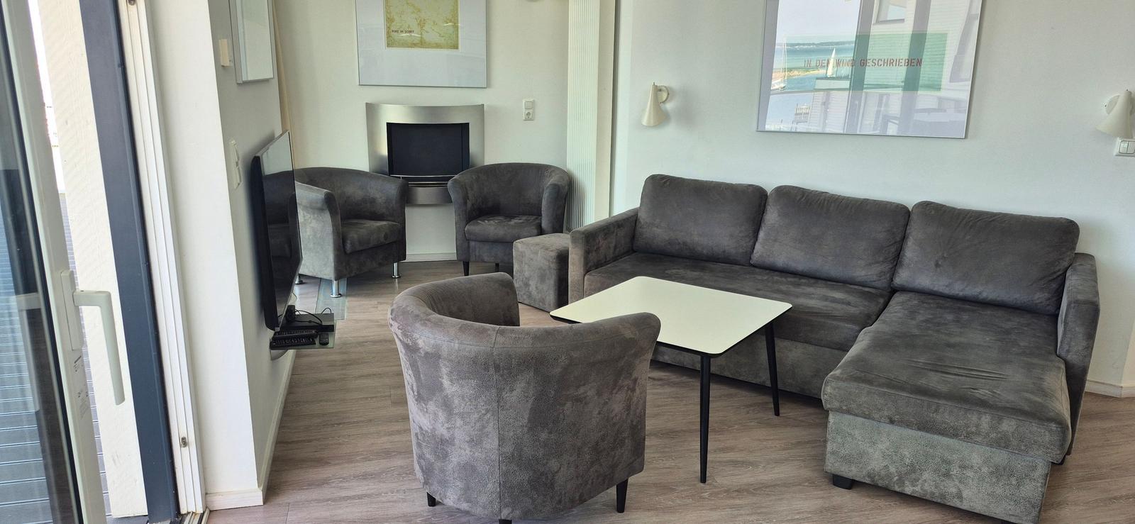 Wohnzimmer mit grauem Sofa, zwei Sesseln und Flachbild-TV an der Wand.