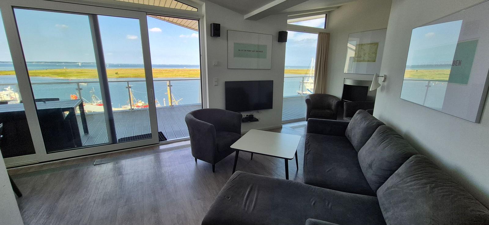 Wohnzimmer mit Sofa, Sessel und TV. Große Fenster mit Blick auf Hafen und Wasser.