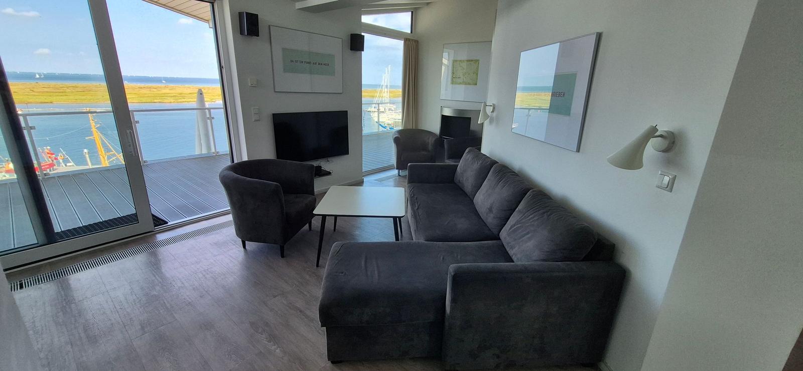 Wohnzimmer mit Sofa, Sessel und TV. Balkon mit Blick auf Wasser und Schiffe.
