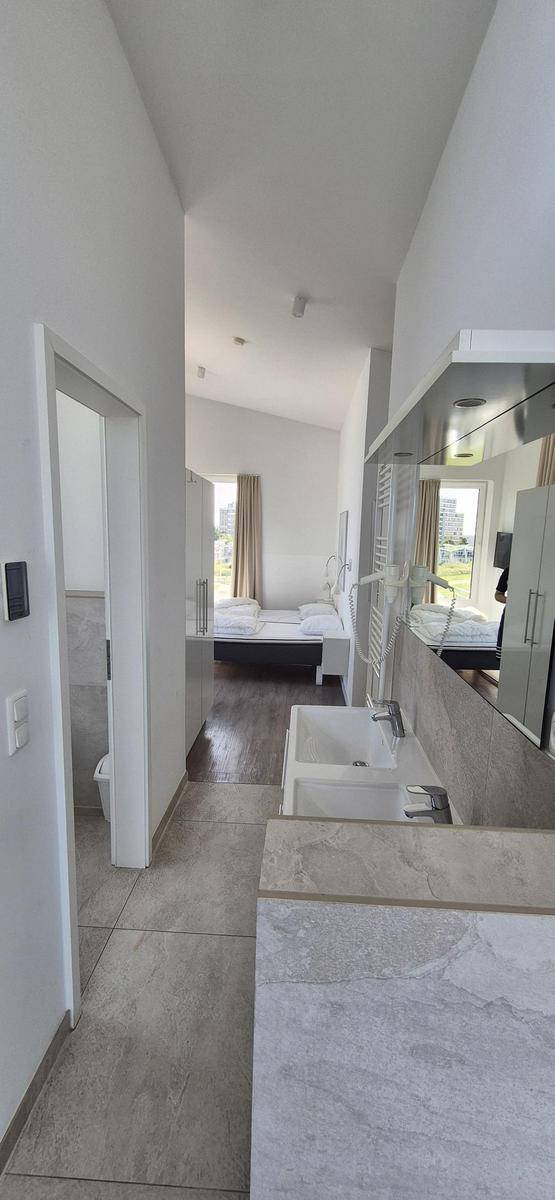 Badezimmer mit Waschbecken und Spiegel. Blick in das Schlafzimmer mit Bett und Fenster.