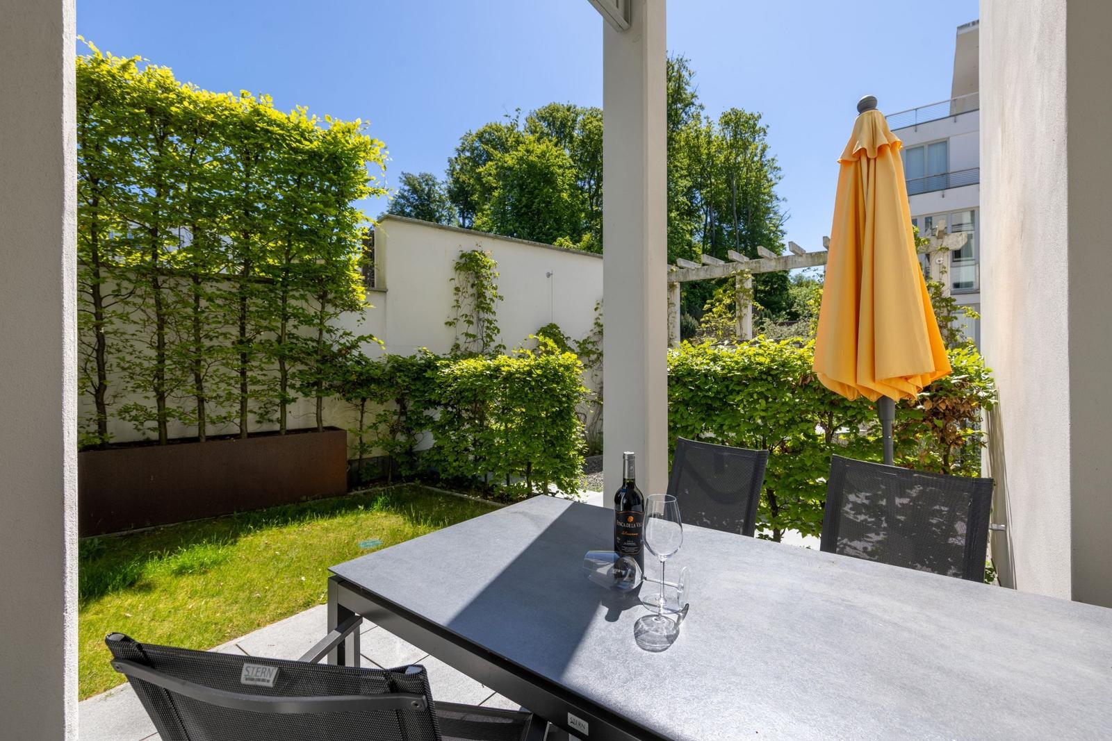 Terrasse mit Tisch, Stühlen und gelber Sonnenschirm. Grünfläche und Bäume im Hintergrund.