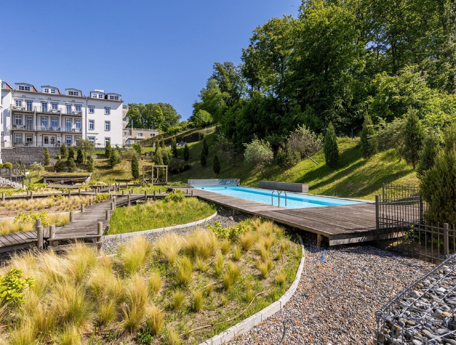 Pool mit Holzterrasse in grüner Umgebung. Im Hintergrund steht ein weißes Gebäude.