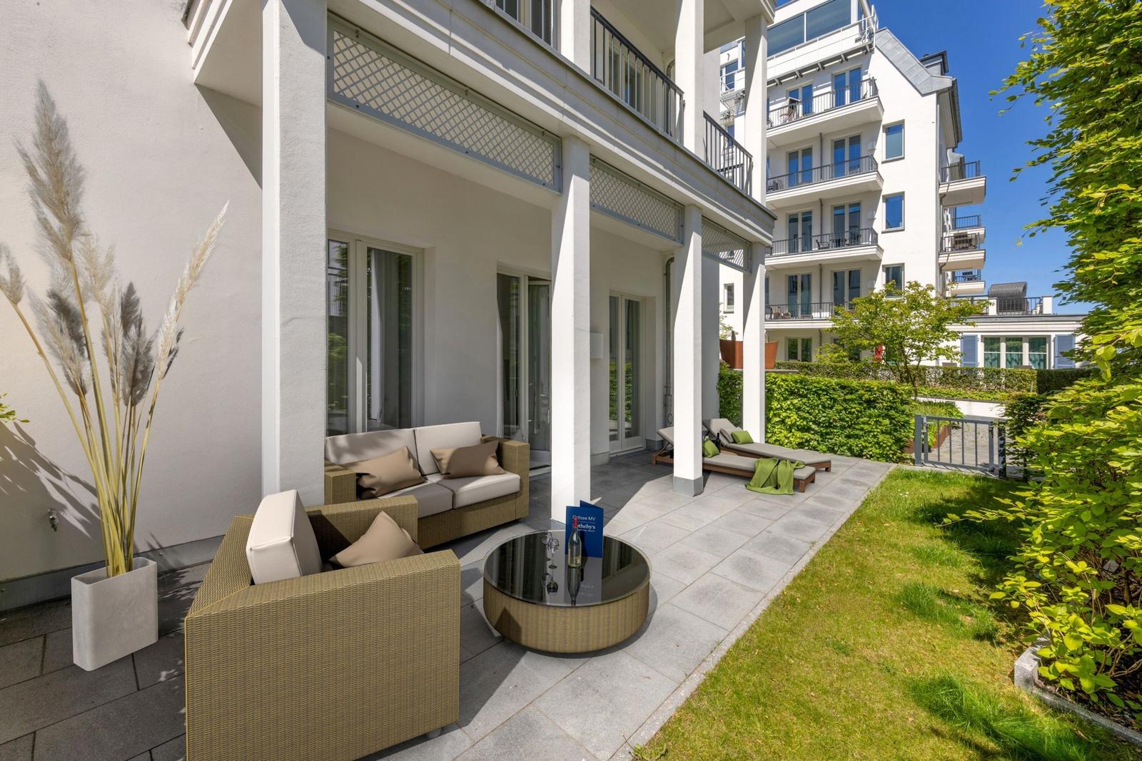 Terrasse mit Sitzgruppe, Liegen und Grünfläche. Modernes Gebäude im Hintergrund.