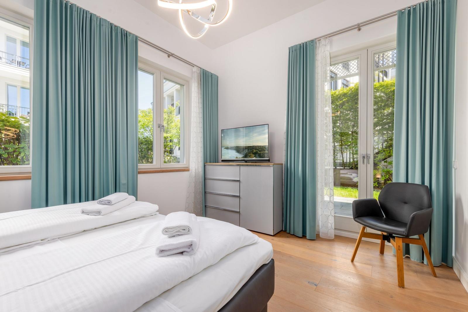 Schlafzimmer mit Bett, TV, Stuhl und Fenstern mit Blick auf Grün.