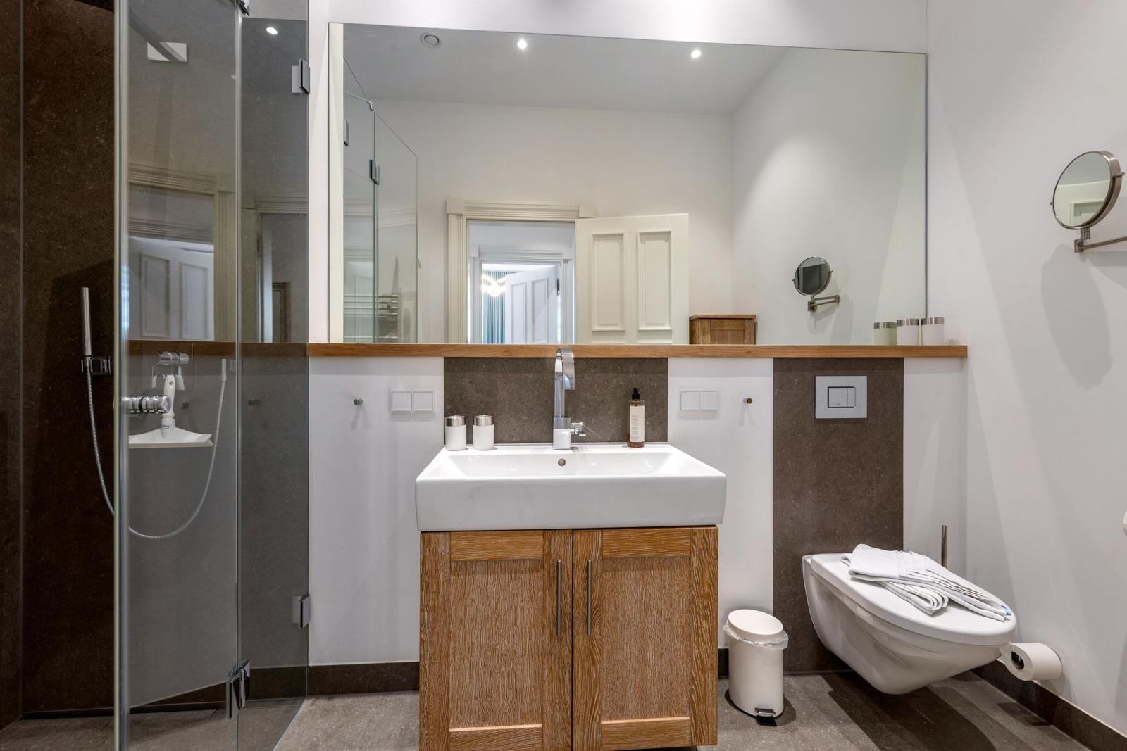 Badezimmer mit Dusche, Waschbecken und WC. Holzunterschrank und großer Spiegel.