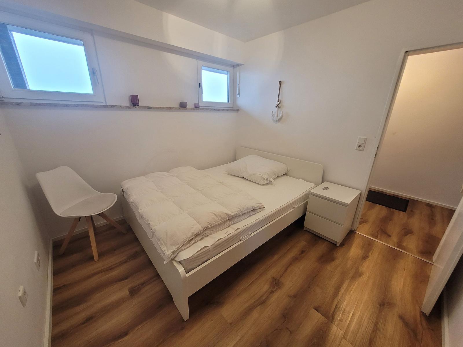 Schlafzimmer mit weißem Bett, Nachttisch und Stuhl. Holzboden und Fenster.