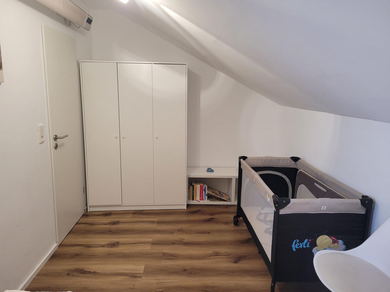 Kammer mit Babybett, Kleiderschrank und Holzboden unter Dachschrägen.