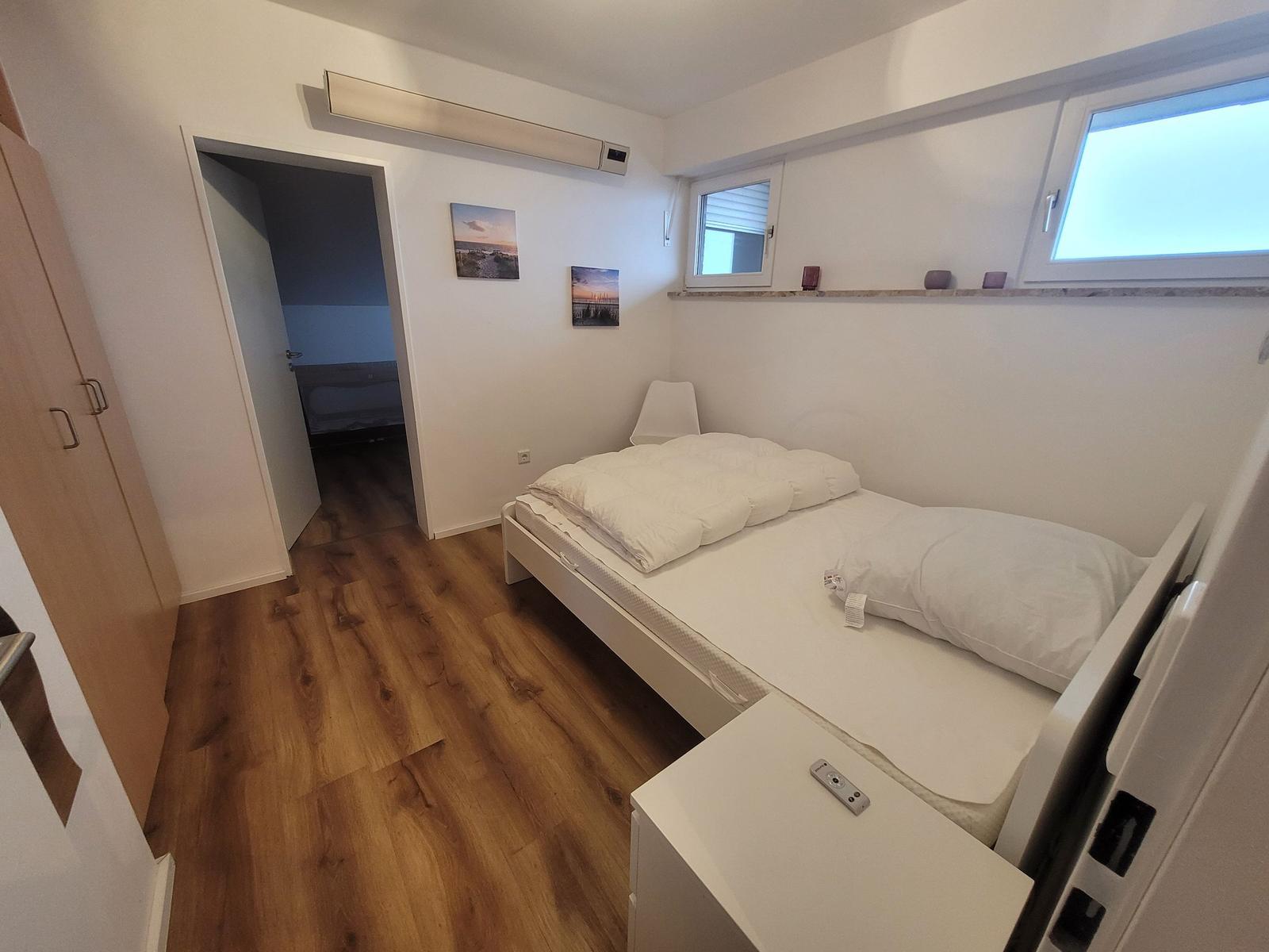 Schlafzimmer mit Bett, Kleiderschrank und Fenster. Tür führt in Bad.